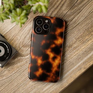 以下が含まれることがあります： 茶色とオレンジの色合いのべっ甲模様のスマートフォンケース。ケースは光沢仕上げで、黒い縁取りがあります。携帯電話ケースは木製の表面にあります。
