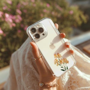 May include: Clear phone case with a floral design featuring yellow and orange flowers. The case is held in a hand, showcasing the phone's camera and Apple logo. The background is blurred with pink flowers.