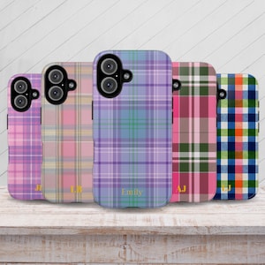 May include: Five phone cases with plaid patterns in pink, peach, purple, green, and blue. Each case has a camera cutout and a personalized initial or name in a script font. The cases are displayed on a light wood surface.