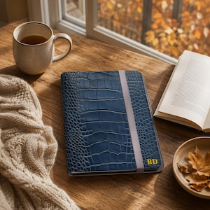 Pode incluir: Uma capa retangular para tablet em azul escuro com um padrão texturizado de pele de crocodilo e uma fita elástica vertical cinza-claro. O canto inferior direito apresenta as letras BD douradas gravadas na superfície azul.