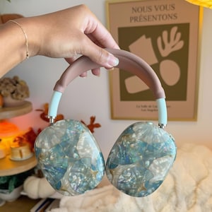 May include: A pair of over-ear headphones with a light blue and iridescent shell design. The headband is a muted pink, and the ear cup arms are light blue. The headphones are held by a person's hand.