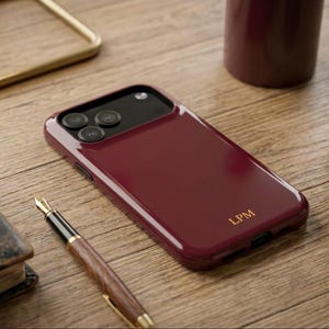 May include: A deep burgundy smartphone case with a glossy finish and the gold-colored initials LPM printed on the lower right corner. The case features a black camera module cutout with three circular lens openings on the upper left side.