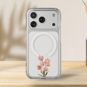 Könnte beinhalten: Transparente Handyhülle mit einem Blumendesign aus rosa und pfirsichfarbenen Blüten. Die Hülle hat einen weißen, kreisförmigen magnetischen Ladebereich. Die Handyhülle liegt auf einer Holzoberfläche.
