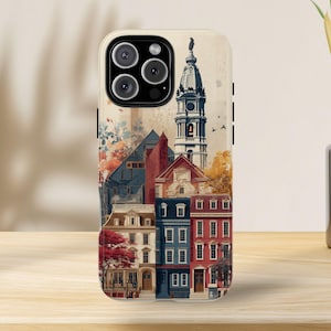 以下が含まれることがあります： カラフルな都市景観のイラストが描かれた携帯電話ケース。赤、青、ベージュの色合いの建物が特徴で、高い尖塔と秋の紅葉が描かれています。ヴィンテージ感のあるデザインで、スマートフォン用です。