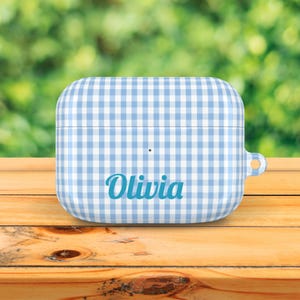 May include: A blue and white gingham patterned case for wireless earbuds. The case has the name "Olivia" printed in a matching blue script font. The case is sitting on a wooden surface.