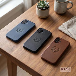 Puede incluir: Tres fundas rectangulares para smartphone en azul marino, negro y marrón, cada una con un emblema de corona de laurel grabado con el texto Custom Logo en el centro. Las fundas tienen un recorte para cámara de doble lente y acabado mate.