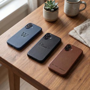 Puede incluir: Tres fundas rectangulares para smartphone de material similar al cuero en azul marino, negro y marrón. La funda azul marino lleva el nombre William, la negra muestra el nombre Olivia con un corazón y la marrón la fecha en números romanos XII-XXIV-MMXXII.