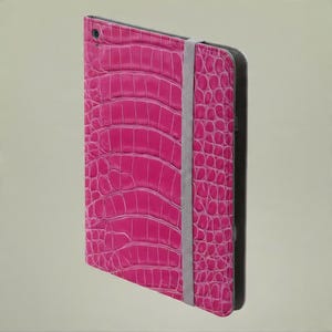 May include: A bright pink tablet case featuring a textured crocodile skin pattern. The rectangular cover includes a vertical grey elastic strap positioned along the right side to secure the device, with a small circular cutout at the top corner.
