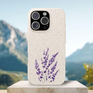 Könnte beinhalten: Eine beige, gesprenkelte Handyhülle mit einem aufgedruckten Motiv aus violetten Lavendelzweigen. Die Hülle verfügt über einen rechteckigen Ausschnitt für ein Dreifach-Kamerasystem und einen Blitz oben links sowie eine matte, texturierte Oberfläche.