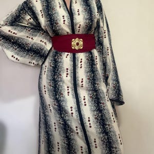May include: A patterned robe in cream, navy, and burgundy. The robe has a vertical stripe design with small burgundy squares. A wide, burgundy belt with a gold-coloured buckle cinches the waist, creating a defined silhouette.