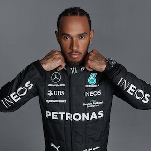 Puede incluir: Un mono de carreras negro con logotipos blancos, incluyendo "PETRONAS" e "INEOS". La persona lleva un reloj y tiene tatuajes. La persona sostiene el cuello del mono.