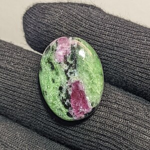 May include: An oval-shaped cabochon gemstone featuring a polished surface with a mix of vibrant green zoisite, deep magenta ruby inclusions, and scattered black hornblende specks, displaying a smooth, rounded, and reflective finish.
