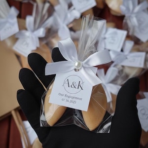 Peut inclure: Deux biscuits chinois dans un sac en cellophane transparent, attachés avec un ruban de satin blanc et une perle. L'étiquette indique "A&K Our Engagement 07.16.2023". Les biscuits sont brun clair. D'autres biscuits emballés sont en arrière-plan.