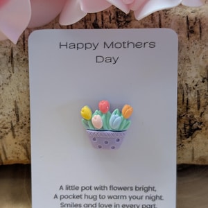以下が含まれることがあります： 「Happy Mothers Day」の文字が入った白いカードと、小さなカラフルな植木鉢のデザイン。 植木鉢には黄色、ピンク、青、オレンジ色のチューリップが入っています。 カードには花と愛についての詩も書かれています。