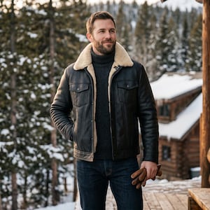 May include: A black leather jacket with a cream shearling collar. The jacket features two chest pockets and quilted sleeves. The person is wearing a dark grey turtleneck sweater and blue jeans. Brown leather gloves are held in the right hand.