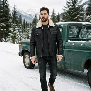 May include: Black leather jacket with a cream-colored shearling collar and cuffs. The jacket has two chest pockets and is worn with a gray sweater and black jeans. The background features a green truck, snow, and a mountain range.