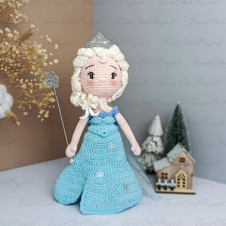 K&ouml;nnte beinhalten: Handgefertigte H&auml;kelpuppe in einem hellblauen Kleid mit Schneeflocken, einer silbernen Krone und einem silbernen Zauberstab. Die Puppe hat blondes Haar und ein rosa Gesicht. Das Thema der Puppe ist eine Prinzessin.