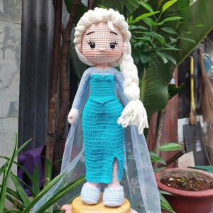 Puede incluir: Mu&ntilde;eca de ganchillo hecha a mano de una princesa con una larga trenza blanca. La mu&ntilde;eca lleva un vestido turquesa con una abertura y una capa transparente. El pelo y el vestido de la mu&ntilde;eca son de hilo. La mu&ntilde;eca est&aacute; sobre una base amarilla.