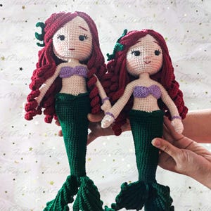 May include: Two handmade crochet mermaid dolls. Each doll has red hair, a purple top, and a green tail. The dolls are holding hands. The dolls are made of yarn and have detailed facial features.