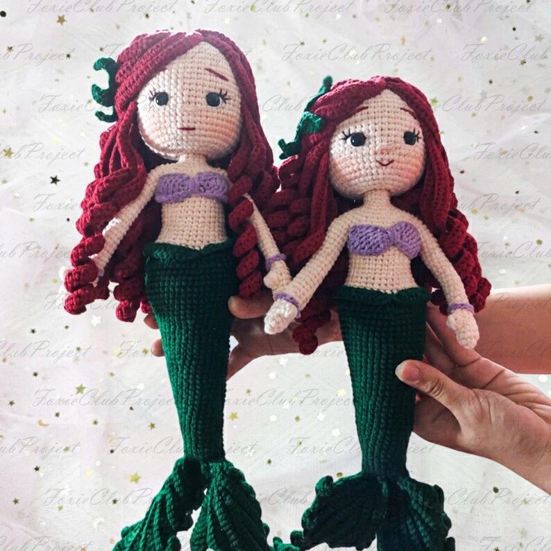 May include: Two handmade crochet mermaid dolls. Each doll has red hair, a purple top, and a green tail. The dolls are holding hands. The dolls are made of yarn and have detailed facial features.