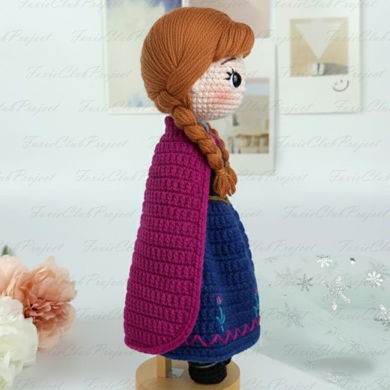 May include: A handmade crochet doll featuring a character with auburn hair in braids, a pink cape, and a blue dress with embroidered details. The doll is displayed on a wooden stand, showcasing the intricate crochet work.
