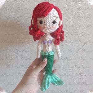 May include: Crocheted mermaid doll with red hair, a white face, and a green tail. The doll has a purple top and a light green skirt. The doll is handmade and has a whimsical design.