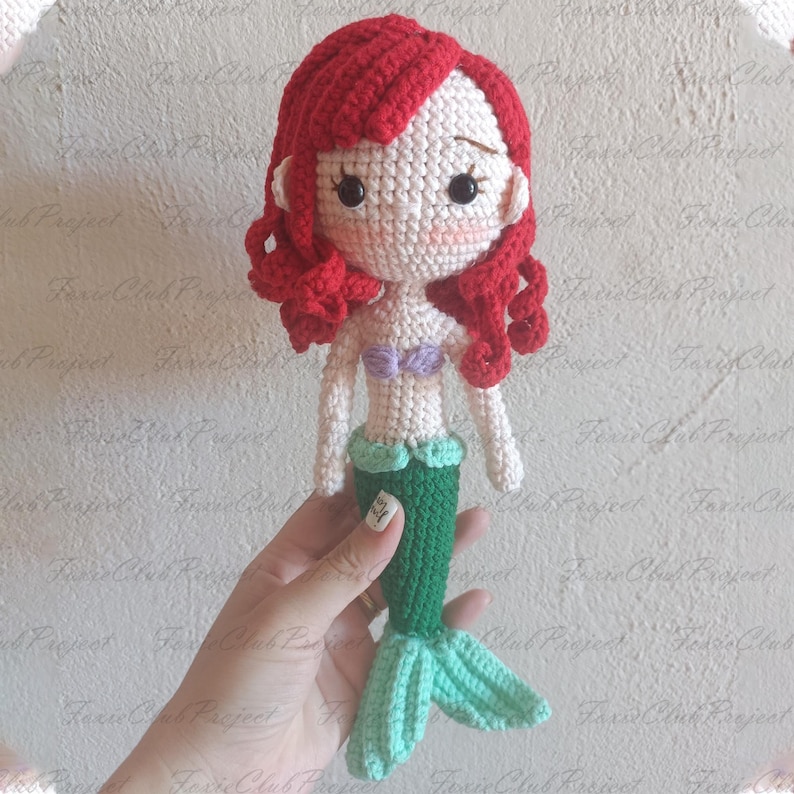 May include: Crocheted mermaid doll with red hair, a white face, and a green tail. The doll has a purple top and a light green skirt. The doll is handmade and has a whimsical design.