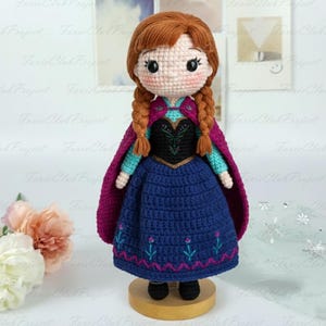 May include: A handmade crochet doll featuring a character with auburn braids, wearing a blue dress with floral embroidery, a black bodice, and a magenta cape. The doll is displayed on a wooden stand, showcasing intricate details.