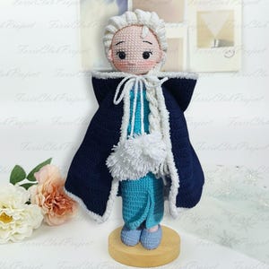 Puede incluir: Mu&ntilde;eca de ganchillo hecha a mano que representa a un personaje con piel clara, cabello blanco y mejillas rosadas. La mu&ntilde;eca viste un conjunto azul y blanco con una capa azul oscuro. La mu&ntilde;eca est&aacute; sobre una base de madera, mostrando un dise&ntilde;o artesanal caprichoso.