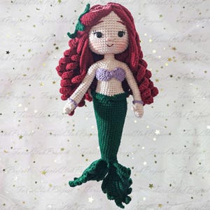May include: A handmade crochet mermaid doll with red hair, a purple seashell top, and a green tail. The doll has a smiling face and is wearing purple bracelets. The mermaid is a whimsical, handcrafted item.