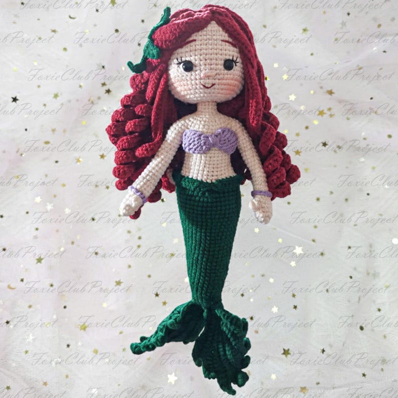 May include: A handmade crochet mermaid doll with red hair, a purple seashell top, and a green tail. The doll has a smiling face and is wearing purple bracelets. The mermaid is a whimsical, handcrafted item.