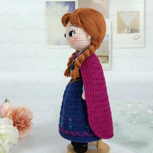 May include: A handmade crochet doll featuring a character with auburn hair in braids, wearing a blue dress with floral embroidery and a magenta cape. The doll is standing on a wooden stand, showcasing the intricate details of the craft.