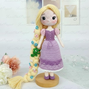 Könnte beinhalten: Handgefertigte Häkelpuppe einer Prinzessin mit langem blondem Haar, verziert mit blauen und rosa Blumen und einem grünen Frosch. Die Puppe trägt ein lila Kleid mit rosa Ärmeln und lila Schuhen. Die Puppe steht auf einem Holzsockel.