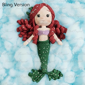 May include: A handmade crochet mermaid doll with red hair, a purple top, and a green tail. The doll has white arms and is adorned with small embellishments. The background is a light blue with white cloud-like shapes. The text "Bling Version" is in the top left corner.