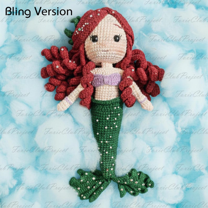 May include: A handmade crochet mermaid doll with red hair, a purple top, and a green tail. The doll has white arms and is adorned with small embellishments. The background is a light blue with white cloud-like shapes. The text "Bling Version" is in the top left corner.