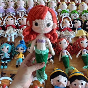 May include: A collection of handmade crochet dolls, including a mermaid doll with red hair and a green tail. Other dolls include characters with red, blonde, and black hair, wearing various outfits. The dolls are displayed on a wooden surface.