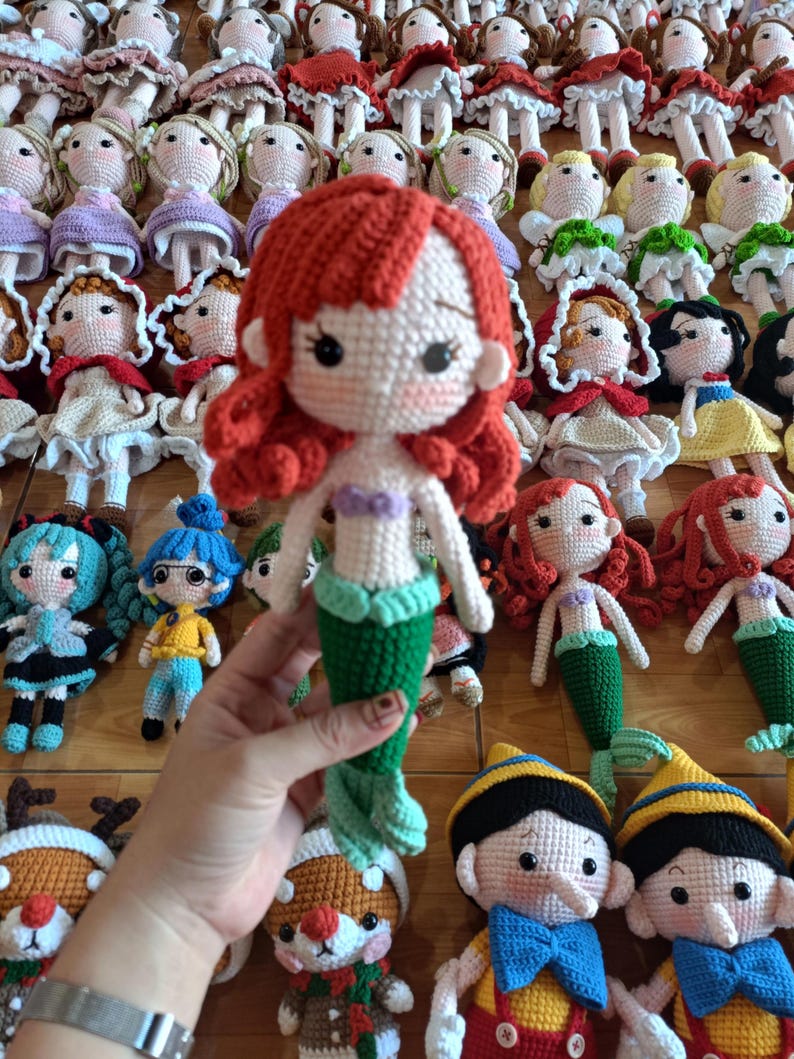 May include: A collection of handmade crochet dolls, including a mermaid doll with red hair and a green tail. Other dolls include characters with red, blonde, and black hair, wearing various outfits. The dolls are displayed on a wooden surface.