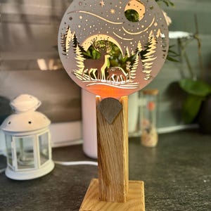 Könnte beinhalten: Dekorative Holzlampe mit kreisförmigem Design, das ein Reh und eine Waldszene darstellt. Die Lampe hat einen Holzsockel und ein Licht, das durch das ausgeschnittene Design scheint. Ein warmer, einladender Akzent.