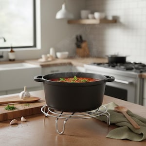 May include: A black cast iron pot filled with tomato soup, garnished with basil, sits on a chrome trivet. Steam rises from the soup, suggesting it is hot. The pot has two handles. A wooden spoon and cutting board are nearby.