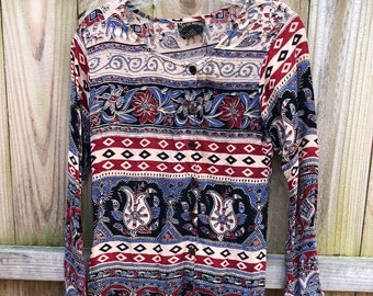 Könnte beinhalten: Langärmlige Tunika mit Knopfleiste, mit einem lebendigen Muster aus Elefanten, Blumen und geometrischen Formen in Rot, Blau, Schwarz und Creme. Die Tunika hängt an einem transparenten Kleiderbügel vor einem Holzzaun.