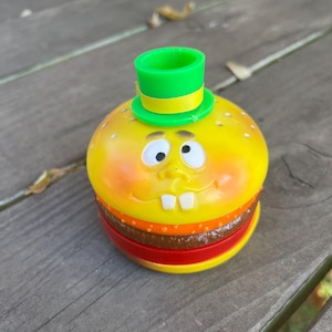 Puede incluir: Una hamburguesa de juguete de plástico amarillo con un sombrero verde y amarillo. La hamburguesa tiene una cara sonriente con dientes blancos y ojos marrones.