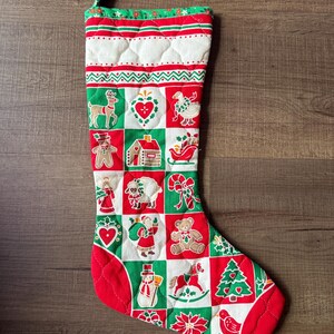 Op de afbeelding: Een gewatteerde kerstsok met een ruitpatroon van rode, witte en groene vierkanten. Elk vierkant toont feestelijke illustraties, waaronder een rendier, peperkoekmannetje, engel, de Kerstman, een sneeuwpop, een teddybeer, een slee en een kerstboom.