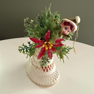 May include: A vintage Christmas bell decoration featuring a green holly-like arrangement with red poinsettias and a small elf figurine peeking out. The bell is white with a decorative openwork design.