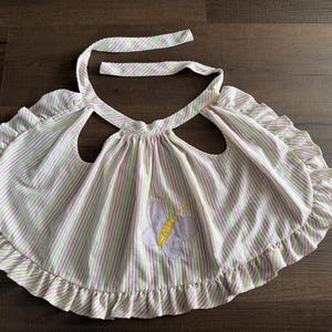 May include: A striped baby bib with a ruffled edge and a butterfly applique. The bib features vertical stripes in pastel colors of pink, yellow, and green. The bib has a tie closure at the neck.