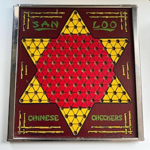 Könnte beinhalten: Ein vintage Spielbrett aus Metall für das Spiel San Loo Chinese Checkers. Das Brett ist rot und gelb mit einem sternförmigen Spielfeld. Das Brett ist in silberfarbenem Metall gerahmt und trägt die Aufschrift "San Loo Chinese Checkers".