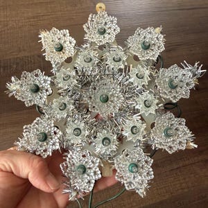 May include: A star-shaped Christmas tree topper featuring a white plastic frame adorned with silver tinsel and numerous clear, textured plastic light covers. The ornament includes multiple small light bulbs and thin green electrical wiring.