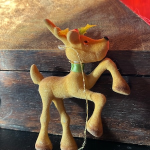 Puede incluir: Un adorno navideño vintage de reno de peluche con una rama de acebo verde y roja en la cabeza, una cinta verde alrededor del cuello y una cuerda dorada para colgar.