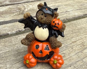 May include: A brown textured bear figurine dressed in a black bat costume with wings, sitting atop a large orange jack-o'-lantern. The bear holds a small orange pumpkin in one arm, and two smaller orange pumpkins are positioned at the base.