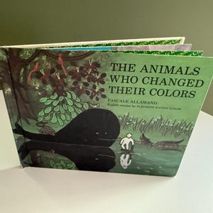 Puede incluir: Un libro infantil de tapa dura titulado The Animals Who Changed Their Colors de Pascale Allamand, versión en inglés de Elizabeth Watson Taylor. La portada presenta un fondo verde con ilustraciones de una gran ballena negra, una tortuga y una pequeña figura blanca de pie cerca del agua, rodeadas de árboles y pájaros.
