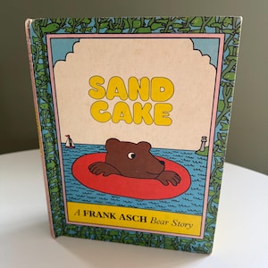 May include: A vintage children's book titled "SAND CAKE" by Frank Asch. The cover features a brown bear in a red inflatable ring in blue water. The book has a green border with a pink and blue background.